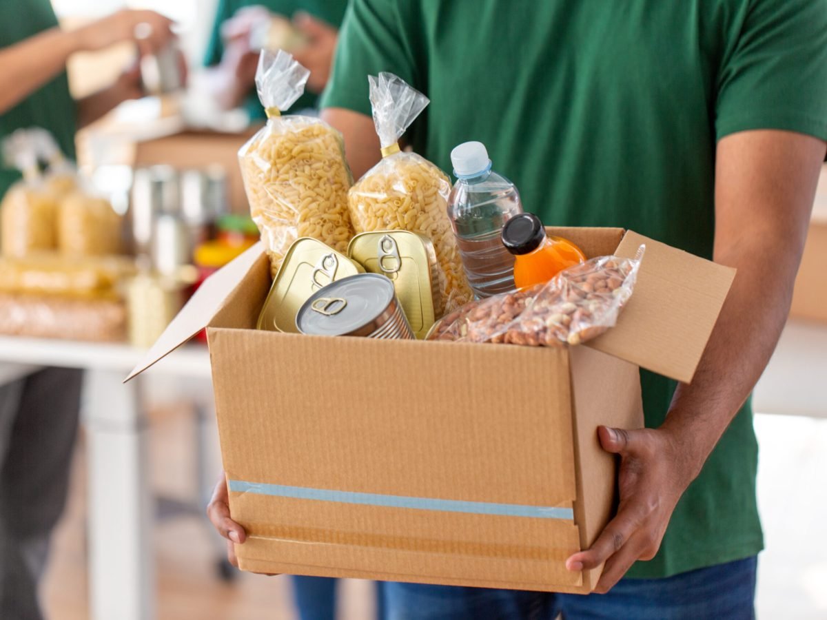 charity, donation and volunteering concept - close up of male volunteer's hands holding box with food over group of people at distribution or refugee assistance center